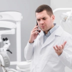 Doctor in white coat talking on a smartphone with a concerned expression in a medical office.